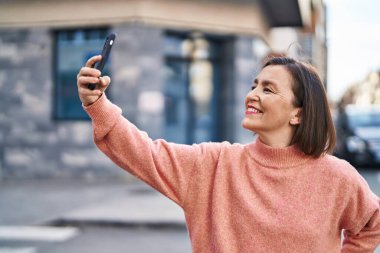 Orta yaşlı kadın gülümsüyor. Sokakta akıllı telefonun yanında özçekim yapıyor.