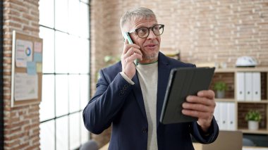 Middle age grey-haired man business worker talking on smartphone using touchpad at office