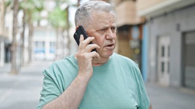 Middle age grey-haired man talking on smartphone with serious expression at street