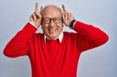 Senior man with grey hair standing over isolated background posing funny and crazy with fingers on head as bunny ears, smiling cheerful 