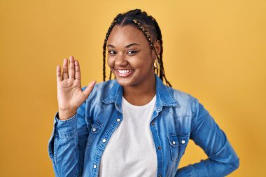 Sarı arka planda dikilmiş örgü ören Afro-Amerikalı kadın merhaba diyor mutlu ve gülümsüyor, dostça karşılama hareketi yapıyor. 