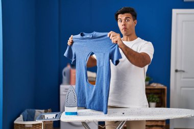 Hispanic man ironing holding burned iron shirt at laundry room making fish face with mouth and squinting eyes, crazy and comical. 