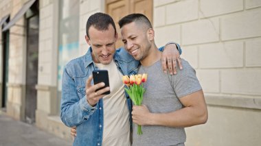 İki adam ellerinde çiçeklerle caddede akıllı telefon kullanıyor.