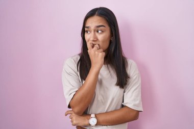Young hispanic woman standing over pink background looking stressed and nervous with hands on mouth biting nails. anxiety problem. 