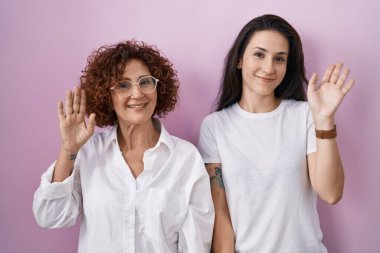 İspanyol anne ve kızı pembe arka plan üzerine gündelik beyaz tişört giyip mutlu ve güler yüzlü, dostça bir jest yapıyorlar. 