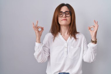 Esmer kadın izole edilmiş beyaz arka planda duruyor rahatlamış ve gülümseyerek gözleri kapalı parmaklarıyla meditasyon hareketi yapıyor. yoga konsepti. 