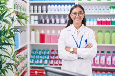 Young beautiful hispanic woman pharmacist smiling confident standing with arms crossed gesture at pharmacy