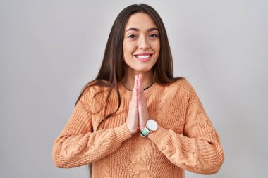 Young brunette woman standing over white background praying with hands together asking for forgiveness smiling confident. 