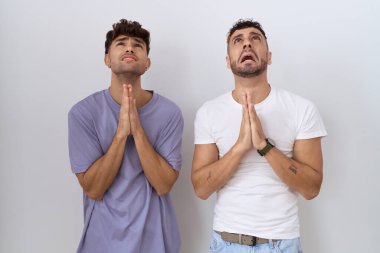 Homoseksüel eşcinsel çift beyaz arka planda durup umutla el ele yalvarıyor ve dua ediyorlar. Yüzlerinde çok duygusal ve endişeli bir ifade var. Yalvarıyor. 