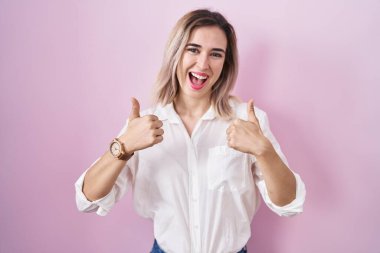 Young beautiful woman standing over pink background success sign doing positive gesture with hand, thumbs up smiling and happy. cheerful expression and winner gesture. 