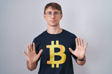 Bitcoin t-shirt giyen beyaz sarışın bir adam. Avuçlarını hareket ettiriyor. Korkmuş ve iğrenç bir ifadeyle reddediyor. Dur ve yasakla. 