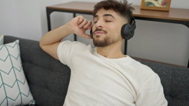 Genç Arap adam evdeki kanepede oturmuş müzik dinliyor.