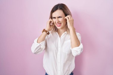 Young beautiful woman standing over pink background covering ears with fingers with annoyed expression for the noise of loud music. deaf concept. 