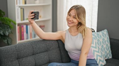 Young blonde woman taking selfie picture with smartphone sitting on the sofa at home