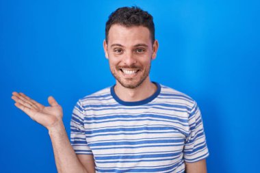 Young hispanic man standing over blue background smiling cheerful presenting and pointing with palm of hand looking at the camera. 