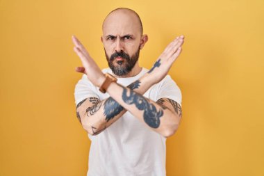 Sarı arka planda duran İspanyol dövmeli genç adam. Reddedilmiş yüz ifadesi. Kollarını kavuşturup negatif işaret veriyor. 