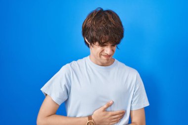 Hispanic young man standing over blue background with hand on stomach because indigestion, painful illness feeling unwell. ache concept. 