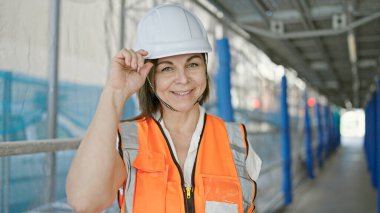 İnşaat alanında elinde kaskla gülümseyen orta yaşlı İspanyol kadın inşaatçı.