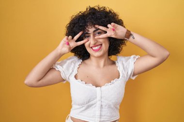 Sarı arka planda duran kıvırcık saçlı genç esmer kadın barış sembolü yapıyor. Yüzünde parmakları, gülümseyerek zaferini gösteriyor. 