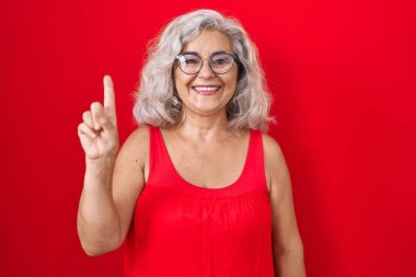 Kırmızı arka planda gri saçlı orta yaşlı bir kadın bir numaralı parmağını gösterip gülümserken kendinden emin ve mutlu.. 