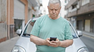 Orta yaşlı, kır saçlı, akıllı telefon kullanan bir adam caddede duruyor.