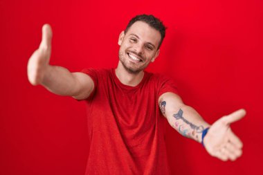 Latin asıllı genç bir adam kırmızı arka planda durup kameraya bakarak sarılmak için kollarını açmış gülümsüyor. Mutluluğu kucaklayan neşeli bir ifade.. 