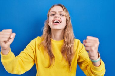 Young caucasian woman standing over blue background angry and mad raising fists frustrated and furious while shouting with anger. rage and aggressive concept. 