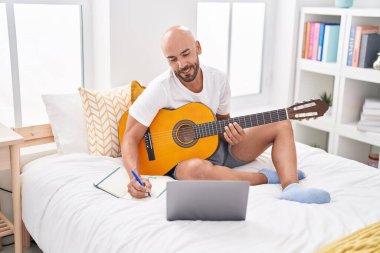 Genç kel adam yatak odasında klasik gitar çalarak şarkı besteliyor.