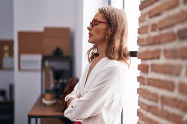 Genç, güzel, İspanyol bir iş kadını. Kollarını kavuşturup ofiste duruyor.