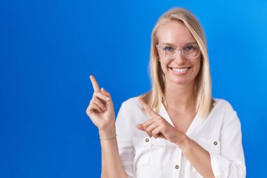 Young caucasian woman standing over blue background smiling and looking at the camera pointing with two hands and fingers to the side. 