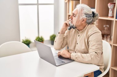 Evdeki masada oturan orta yaşlı, kır saçlı, dizüstü bilgisayar kullanan bir adam.
