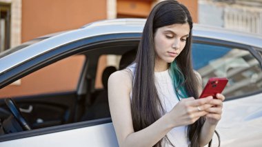 Young beautiful hispanic woman using smartphone leaning on car at street