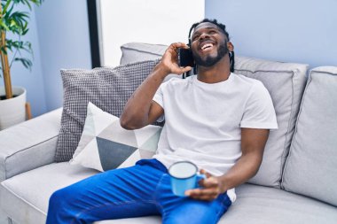 Afro-Amerikalı kadın kahve içiyor ve evde akıllı telefondan konuşuyor.