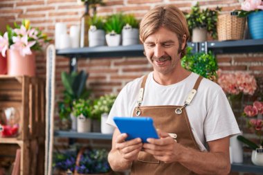 Young blond man florist smiling confident using touchpad at flower shop