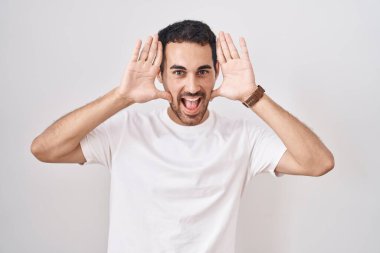 Handsome hispanic man standing over white background smiling cheerful playing peek a boo with hands showing face. surprised and exited 