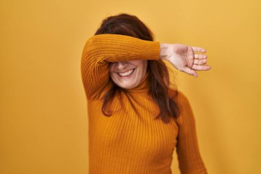 Orta yaşlı İspanyol bir kadın sarı arka planda duruyor. Gözleri kollarıyla kaplıyor. Neşeli ve komik gülümsüyor. kör kavram. 