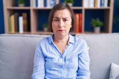 Orta yaşlı İspanyol bir kadın, evde kanepede oturuyor şüpheci ve gergin, sorun yüzünden suratını asıyor. negatif kişi. 