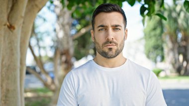 Young hispanic man standing with serious expression at park