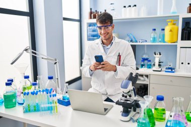 Young hispanic man scientist smiling confident using smartphone at laboratory