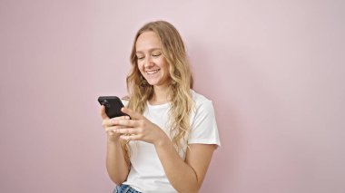 Akıllı telefon kullanan genç sarışın kadın pembe arka planda gülümsüyor.