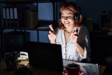 Young caucasian woman working at the office at night gesturing finger crossed smiling with hope and eyes closed. luck and superstitious concept. 