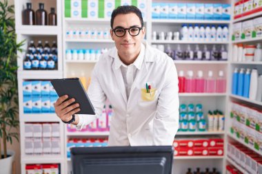 Young hispanic man pharmacist using touchpad and computer at pharmacy