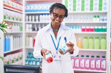 Middle age african american woman pharmacist smiling confident holding medication bottles at pharmacy