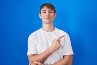 Mavi arka planda duran beyaz tenli sarışın adam, el parmağı yanda, ciddi ve sakin bir yüz ifadesi sergiliyor. 