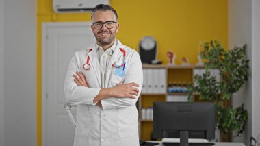 Gri saçlı doktor gülümsüyor. Kollarını kavuşturup klinikte duruyormuş.