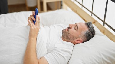 Yatakta uzanmış akıllı telefon kullanan gri saçlı adam.