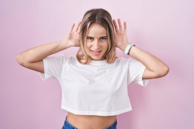 Young blonde woman standing over pink background trying to hear both hands on ear gesture, curious for gossip. hearing problem, deaf 