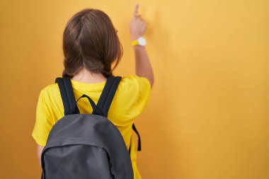 Sarı arka planda öğrenci çantası takan beyaz bir kadın parmak ile öne doğru poz veriyor. 