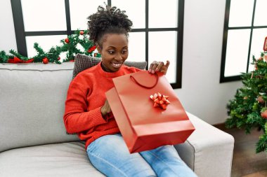 African american woman unpacking christmas gift sitting on sofa at home