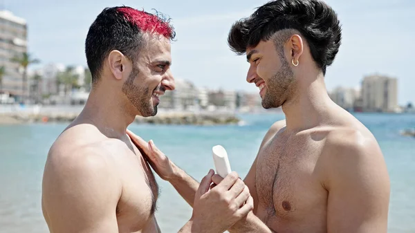 İki erkek turist plajda güneş kremi sürüyor.